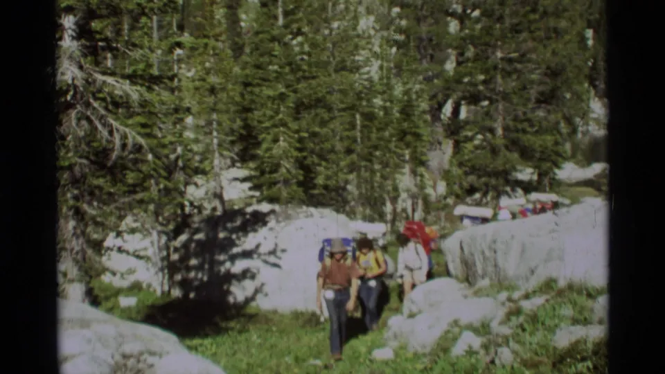1975 Montana home-movie frame showing several backpackers crossing a rocky pine-forest trail with boulders and patchy snow.