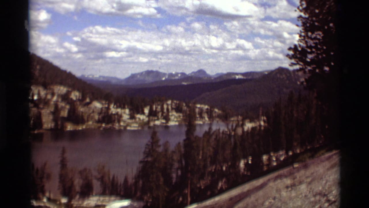 1975 8mm home-movie view of a Montana mountain lake, forested slopes, rocky edges, and distant peaks from a roadside overlook.