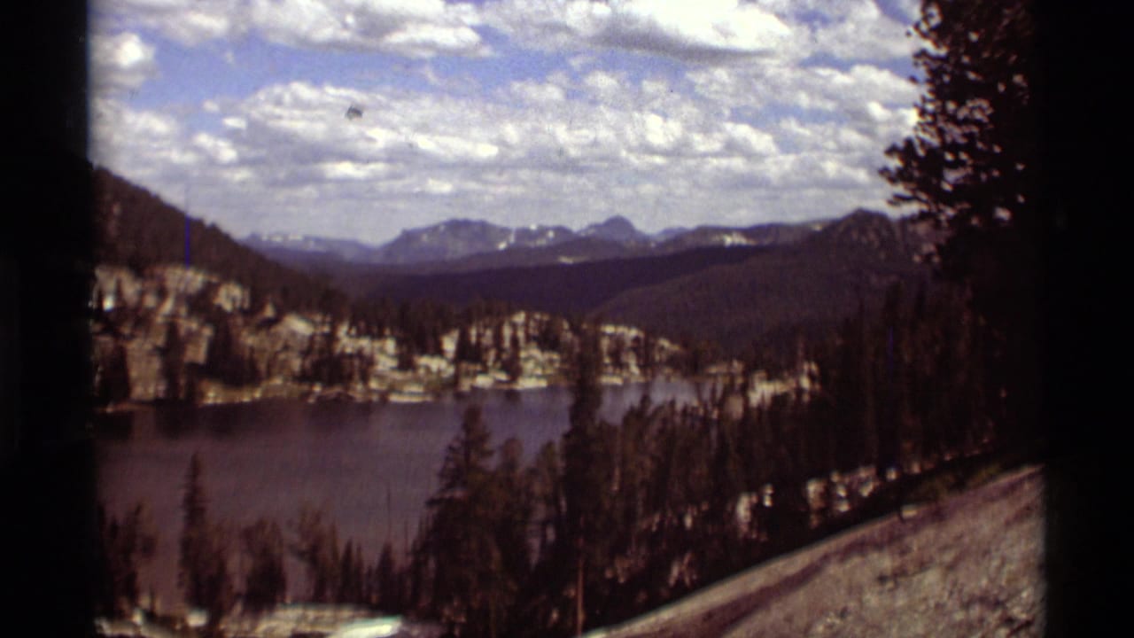 A repeated 1975 Montana scenic view shows the lake, conifers, pale rock faces, and far mountain ridges in soft 8mm color.