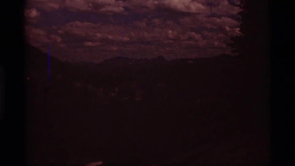 Underexposed 8mm frame of a mountainous landscape, with dark tree and hill forms under a textured cloudy sky.
