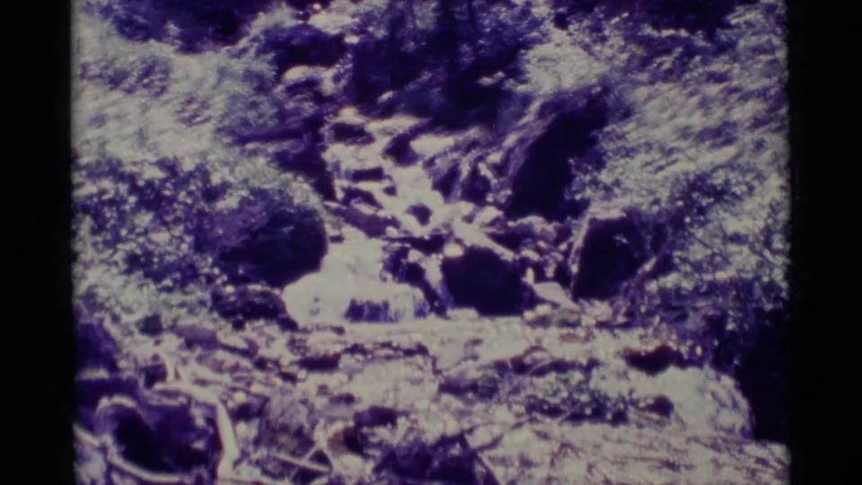 The 1975 home-movie view shows a rocky downhill watercourse surrounded by thick vegetation near Agate Springs, Montana.