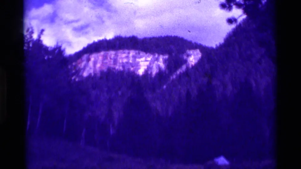 1975 Agate Springs 8mm view of a grassy hillside, distant rocky rise, cloudy sky, and dark tree silhouettes with purple film tint.