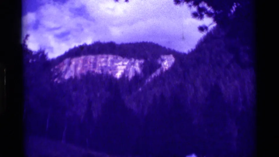 1975 Montana home-movie frame showing a pale cliff face above dense trees, with a small light shape near the lower right.