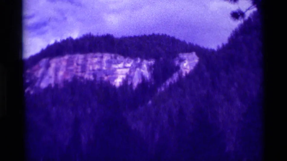 1975 8mm Agate Springs, Montana frame showing a pale rocky cliff rising over dense evergreen trees with strong purple color fading.