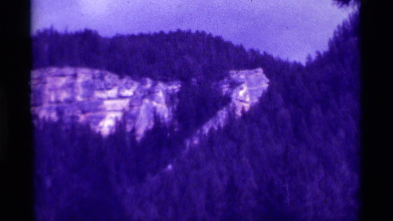 A purple-shifted 1975 8mm view of a wooded Montana ridge with pale exposed rock on the mountainside.