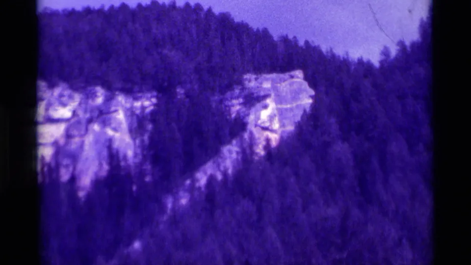 Aged 8mm frame from Agate Springs, Montana, showing a sloping evergreen mountainside with pale rock formations.