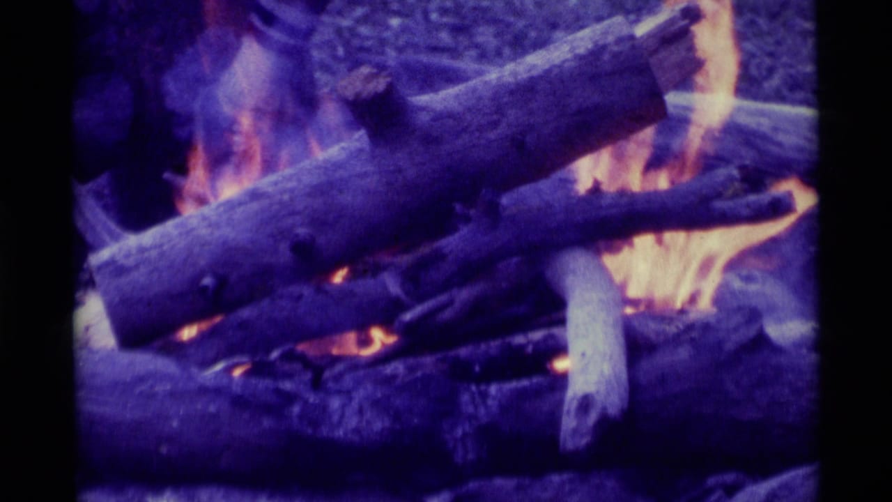 1975 8mm home-movie frame from Agate Springs, Montana, showing logs burning in a campfire with purple-blue film color shift.