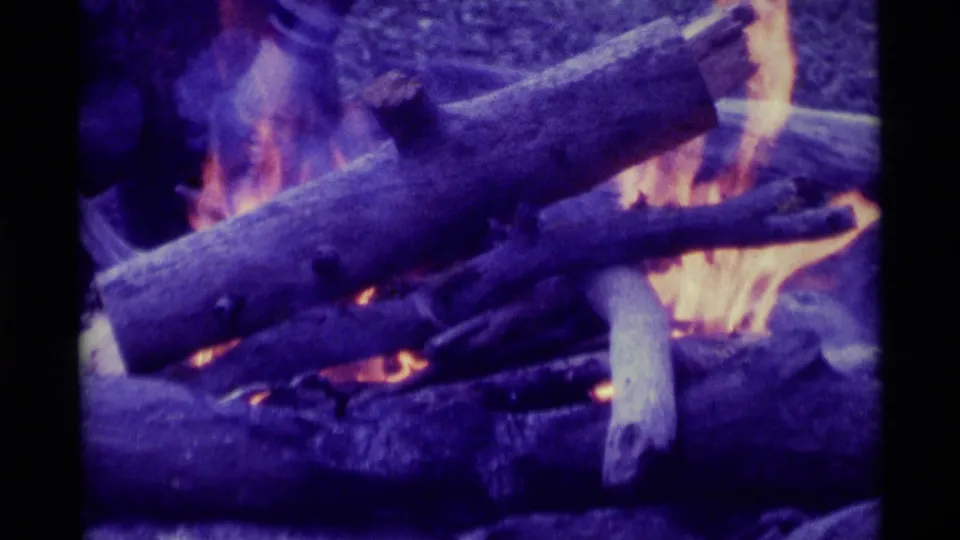 1975 8mm home-movie frame from Agate Springs, Montana, showing logs burning in a campfire with purple-blue film color shift.