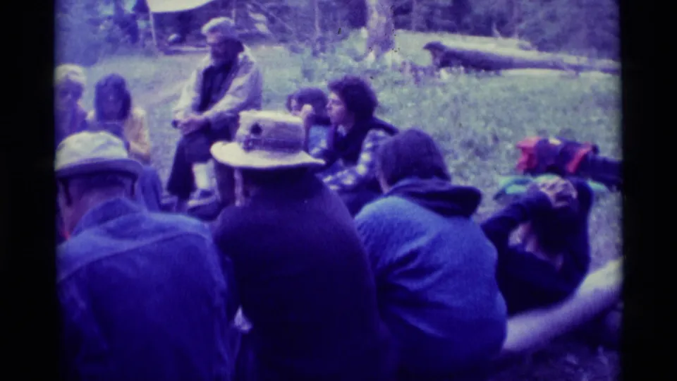1975 Agate Springs home-movie frame showing several adults seated informally on grass around an outdoor campfire gathering.