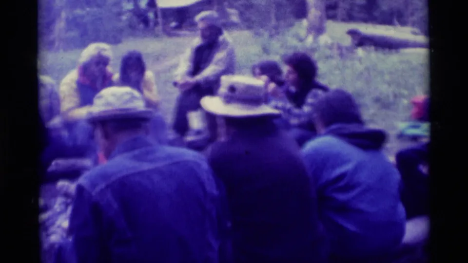 Soft-focus 8mm frame of adults gathered outdoors in Agate Springs, Montana, seated on grass near a campfire in 1975.