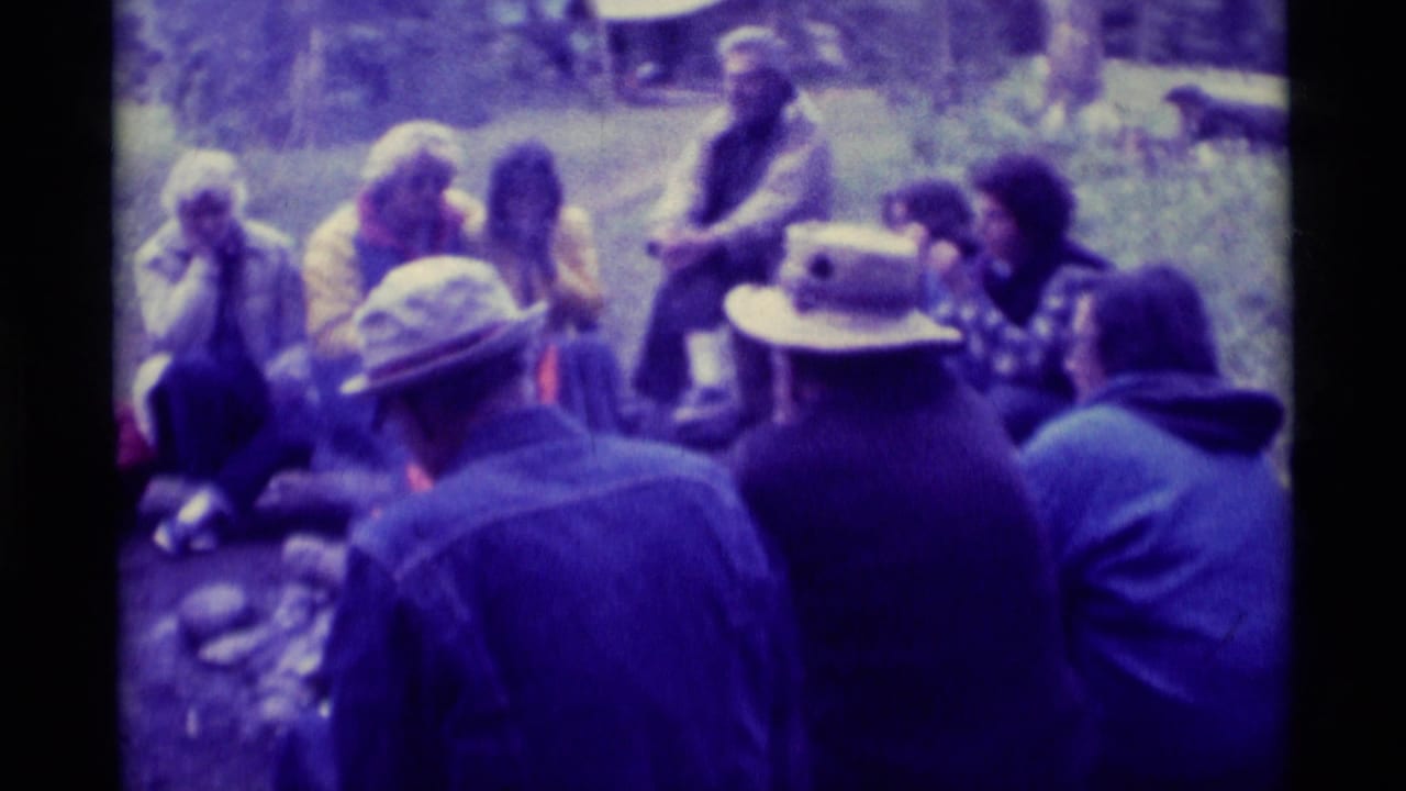 Adults sit in a wooded area around a small campfire, with hats and denim jackets visible.