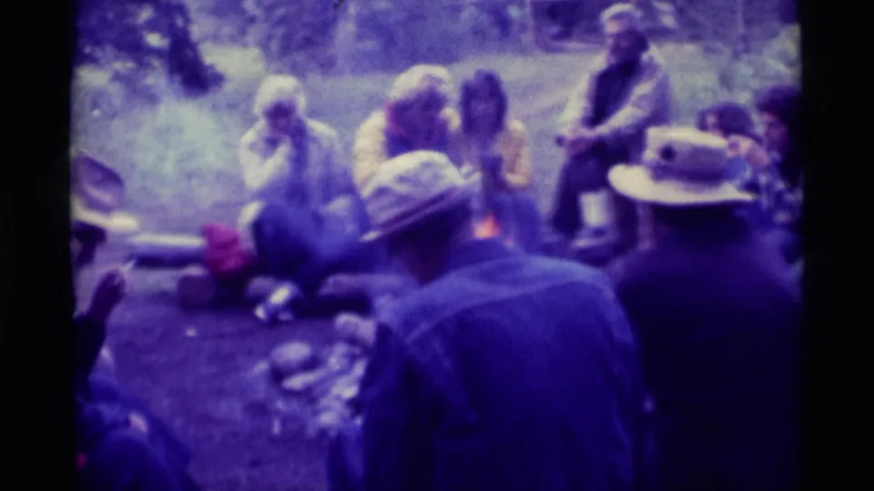 1975 Agate Springs, Montana frame with adults gathered around a campfire, filmed from behind two seated figures in hats.