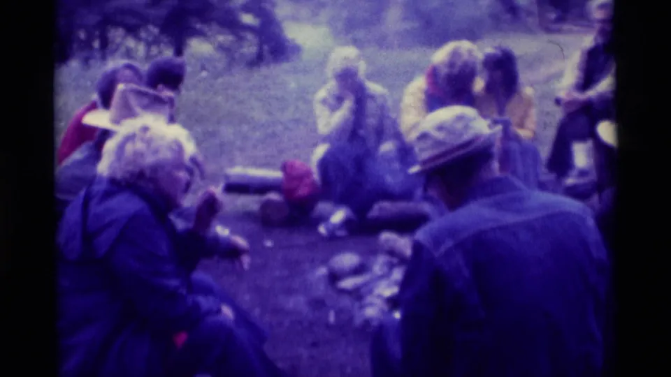 1975 Agate Springs, Montana home-movie frame of adults seated near logs and camp items around an outdoor fire area.