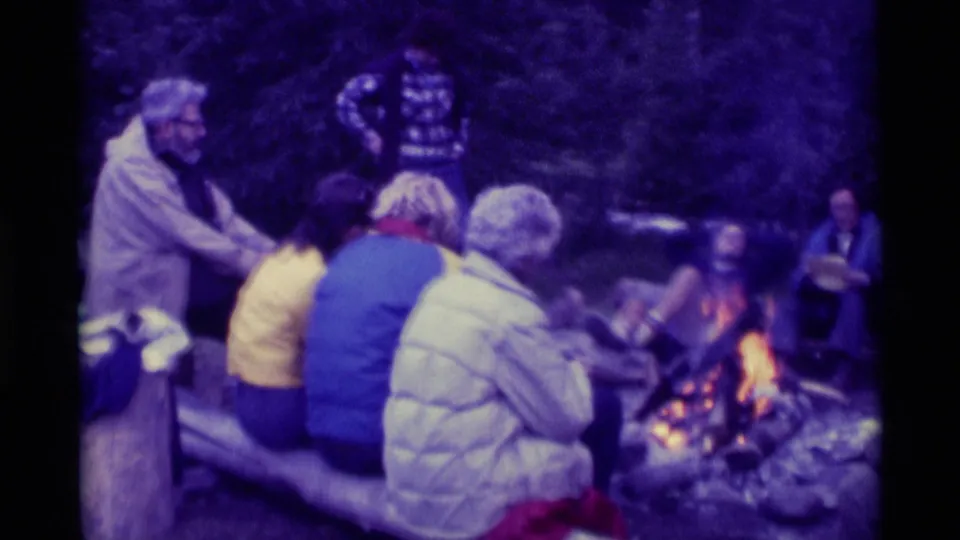 1975 Agate Springs, Montana frame showing adults around a glowing campfire, including seated figures on logs and one standing person.