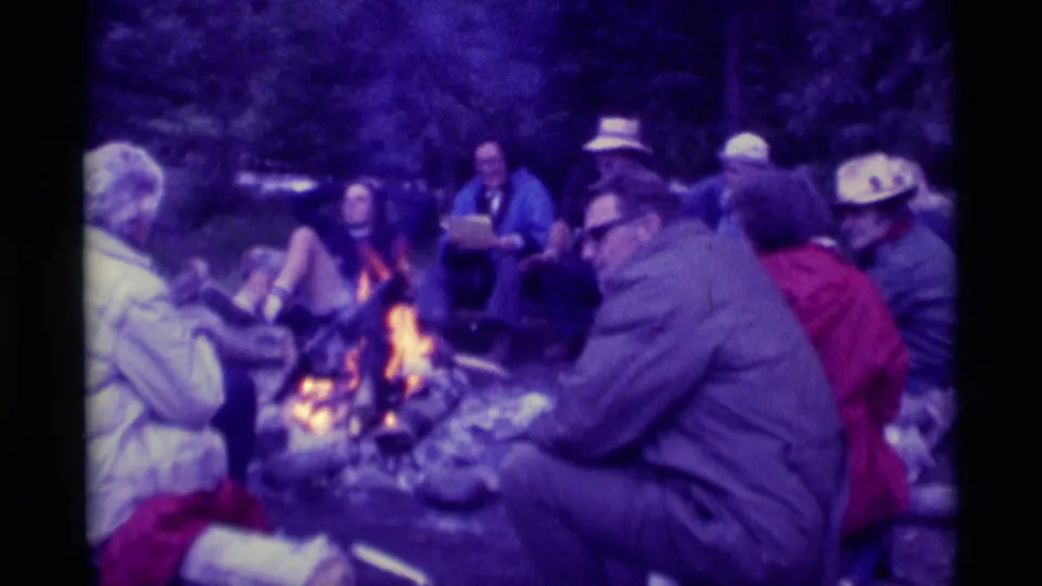 Soft 8mm frame of a campfire gathering in Montana, with seated adults, hats, jackets, and bright firelight in the foreground.
