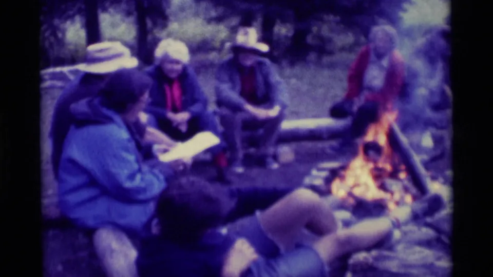 1975 Agate Springs campfire gathering with people seated around logs, one holding papers, and firelight at the edge of frame.