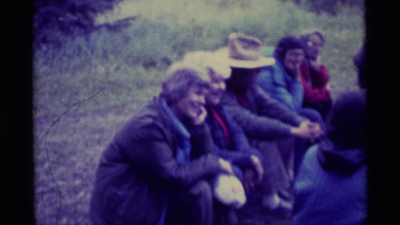 Adults sit close together outdoors in the Montana home-movie frame, with jackets, a brimmed hat, and grassy background visible.