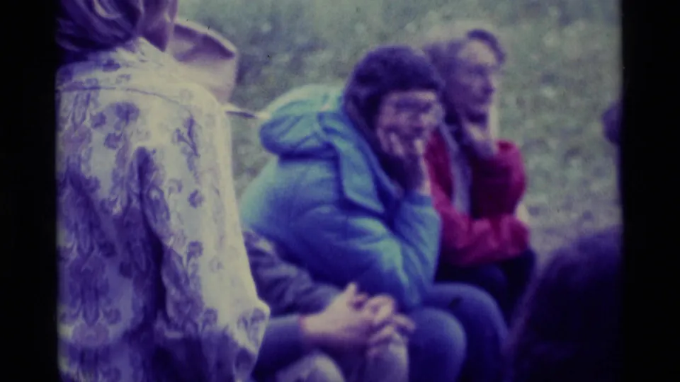 A close 8mm frame shows seated adults at an outdoor Montana rest stop, partly obscured by a patterned garment in the foreground.