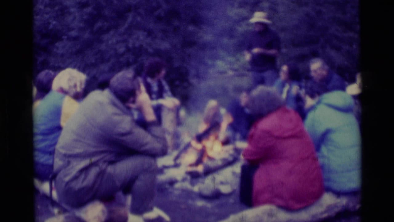 1975 8mm home-movie frame from Agate Springs, Montana, showing a group seated in the woods around a small campfire.