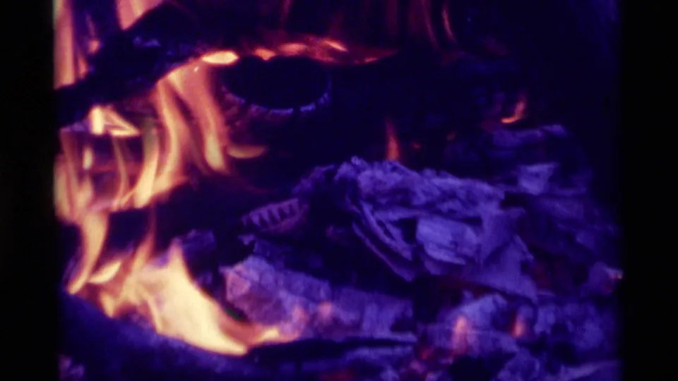 A close 1975 8mm view of campfire flames, charred wood, and glowing embers at the campsite.