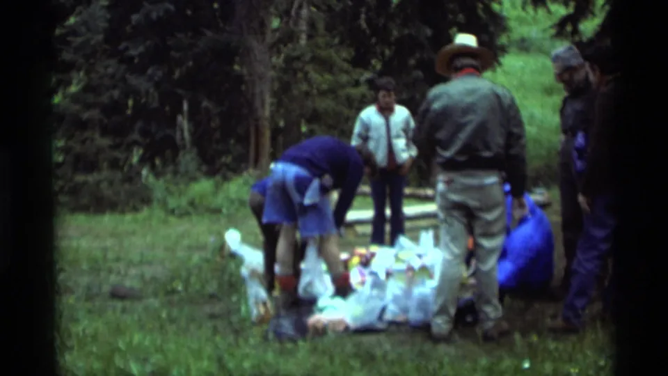 1975 Agate Springs, Montana 8mm frame showing adults around packed supplies in a wooded camping area.