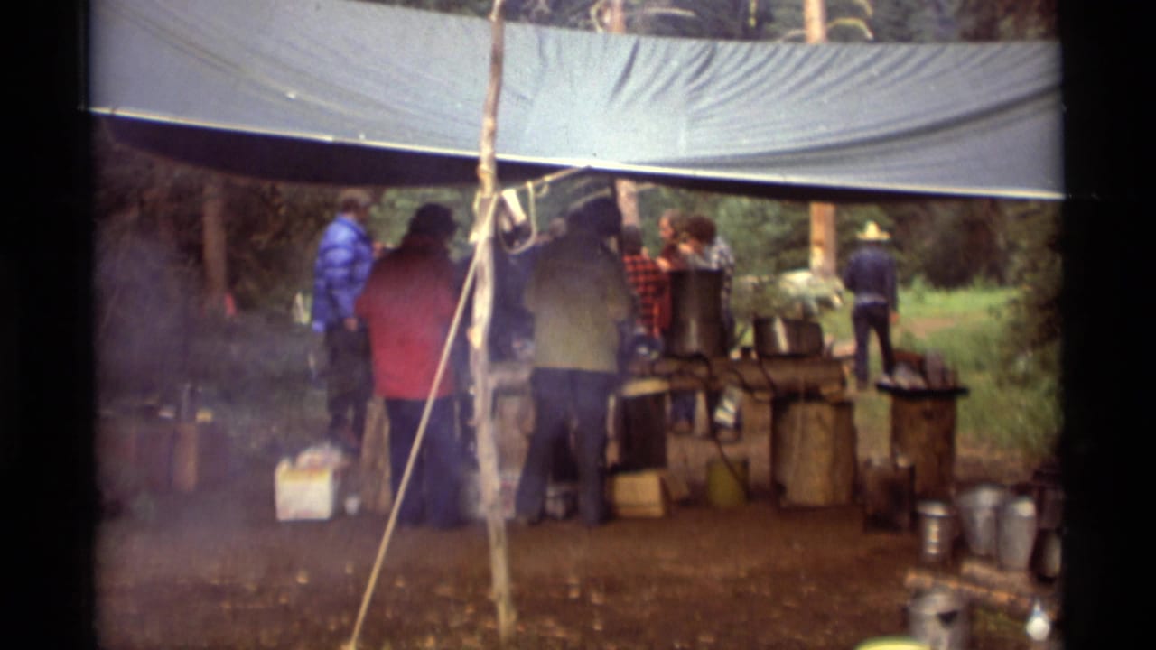 People in jackets gather under a tarp at a wooded outdoor kitchen with smoke and cookware.