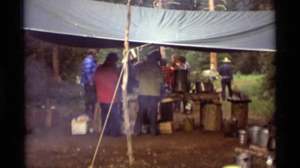 1975 Agate Springs, Montana home-movie frame showing people under a tarp near pots, buckets, supplies, and smoke at a wooded campsite.