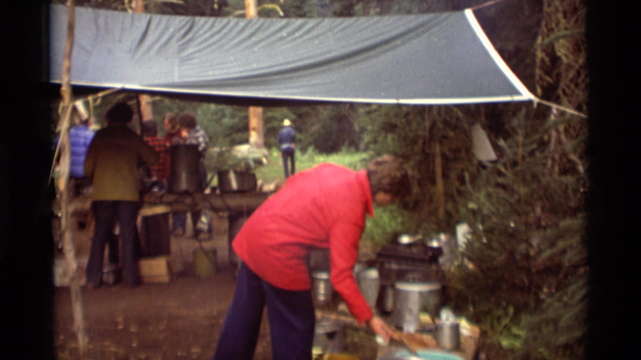 Person in a red jacket bends over cookware beside a tarp-covered camp kitchen.
