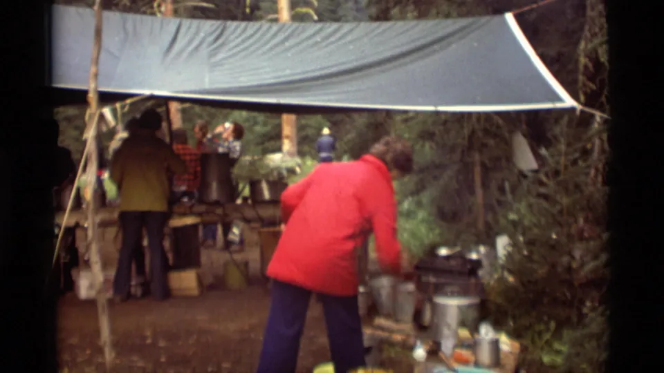 1975 Agate Springs, Montana home-movie frame showing a red-jacketed person working near buckets, pots, and a tarp-covered outdoor kitchen.