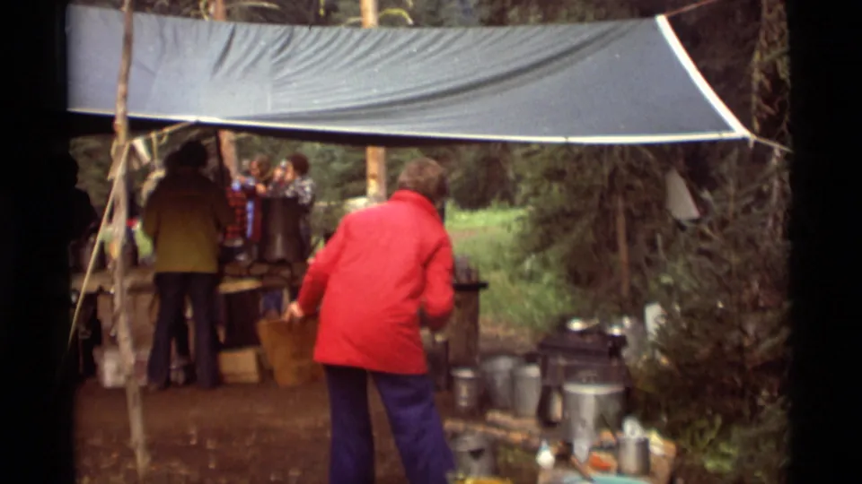 1975 Agate Springs, Montana frame showing a red-jacketed person moving beside camp cookware as others stand at a tarp-covered food area.