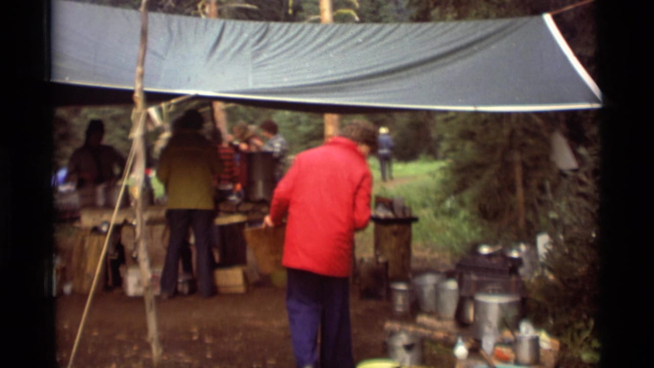 Campers stand beneath a blue tarp near tables with cookware in a wooded camp area.