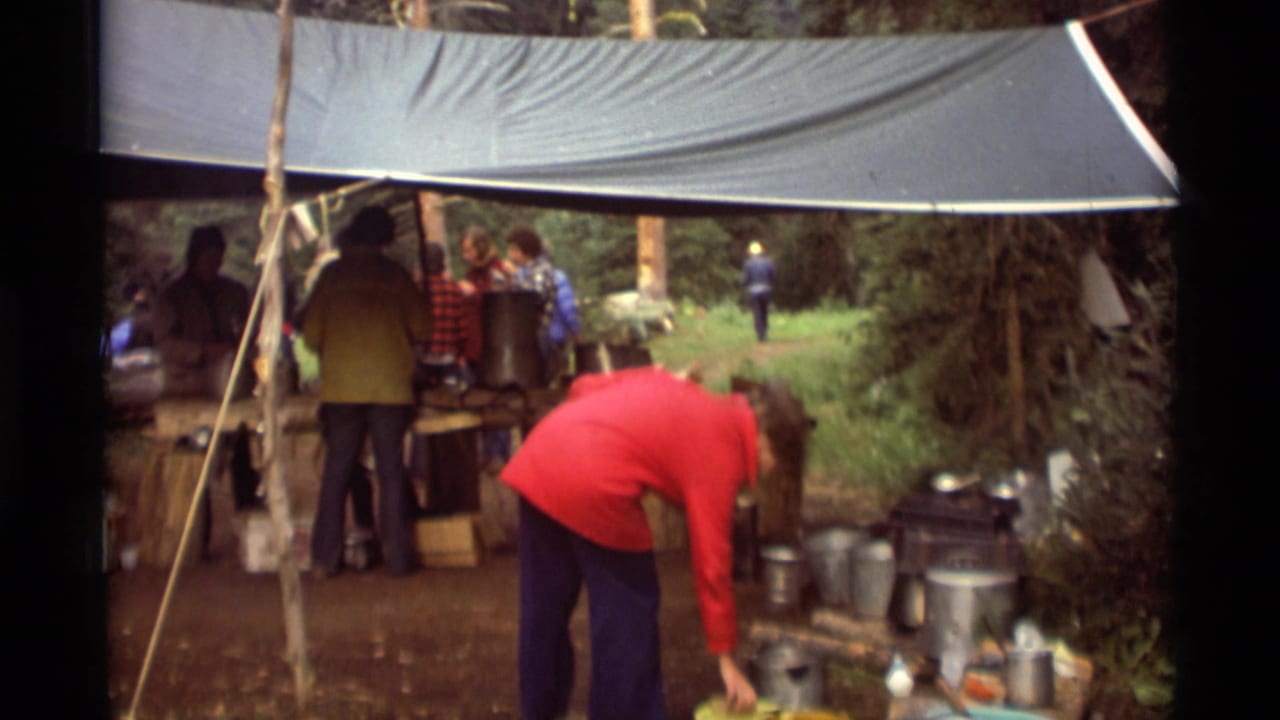 A person in a red jacket bends near cookware while other campers stand under the tarp.