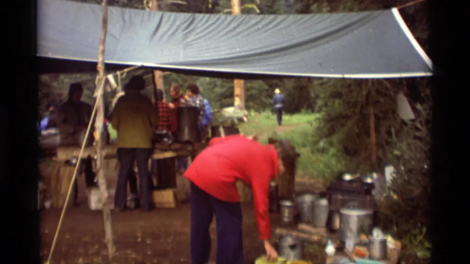 1975 Montana campsite frame with a red-jacketed camper bending beside pots and pans as others work under a tarp.