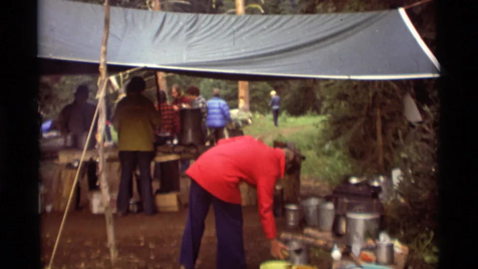 Agate Springs camp frame showing a red-jacketed person reaching near metal pots while campers stand beneath a tarp shelter.