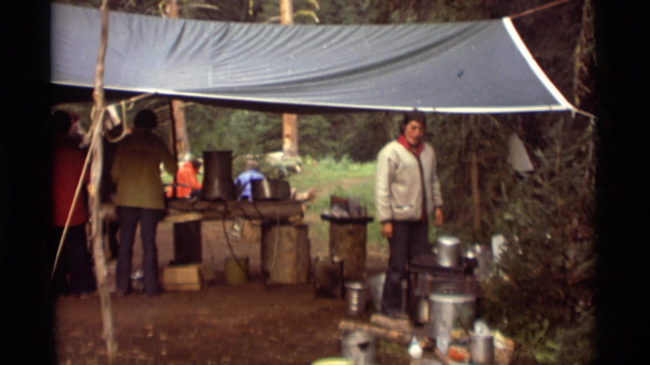 Adults stand under a tarp beside metal pots and a rustic outdoor cooking setup.