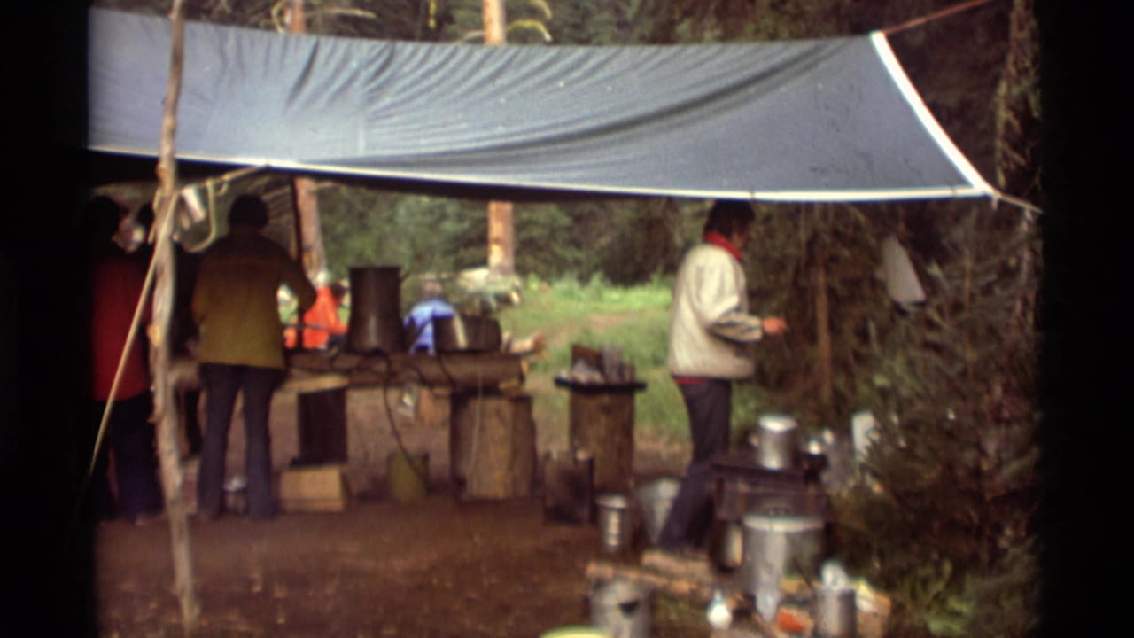 A person in a light jacket moves beside metal pots while others remain under the tarp at the 1975 Montana camp kitchen.