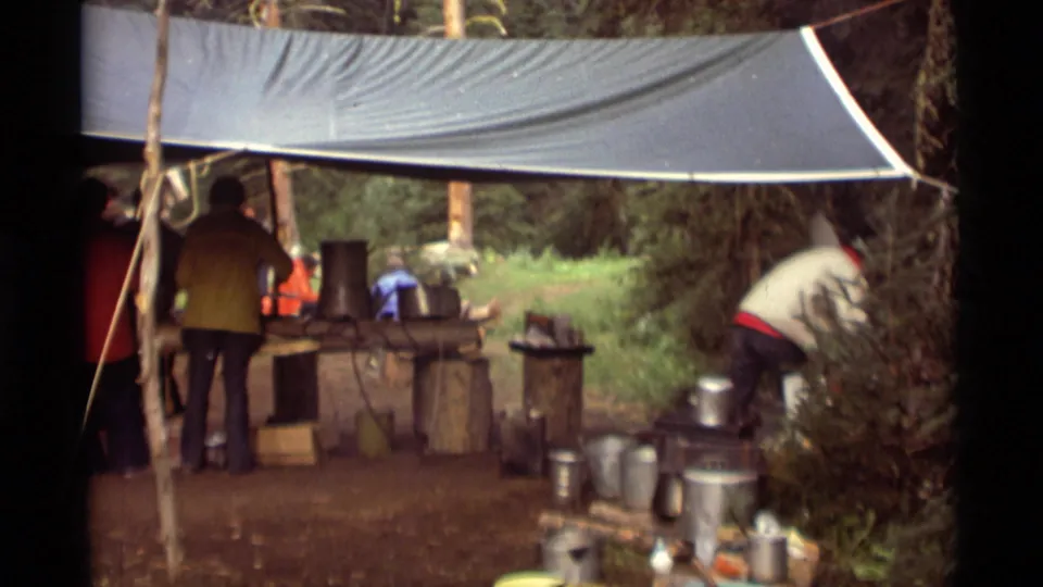 Under the forest tarp, several adults face the camp kitchen while one person bends near pots and containers in the Montana footage.