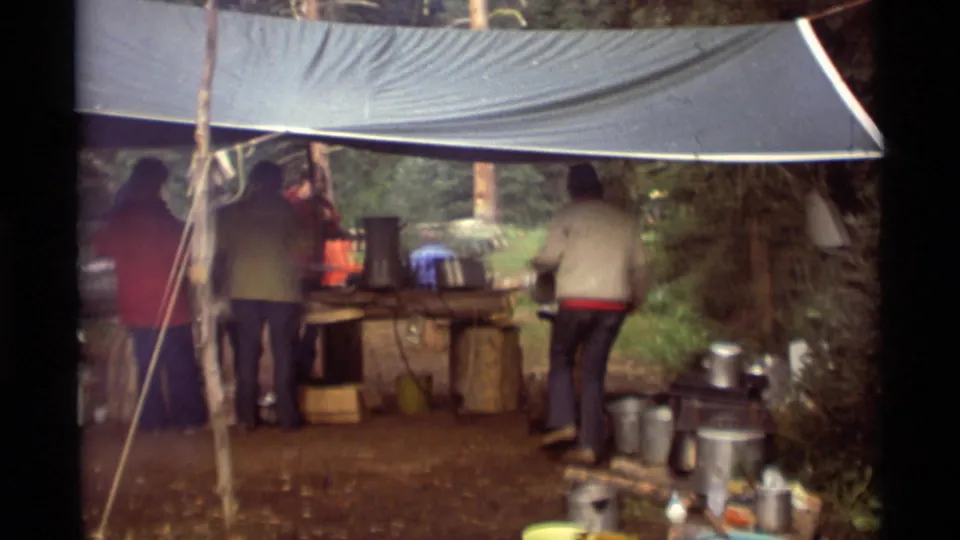 Adults in casual 1970s clothing gather beneath a tarp around a log-supported outdoor kitchen at Agate Springs, Montana.