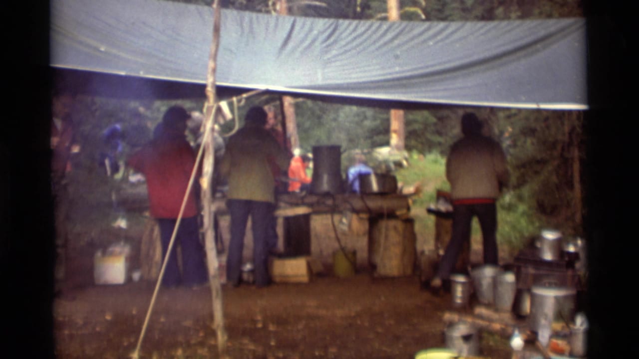 1975 Agate Springs, Montana home-movie frame showing hikers in jackets gathered under a tarp beside cookware and camp supplies.