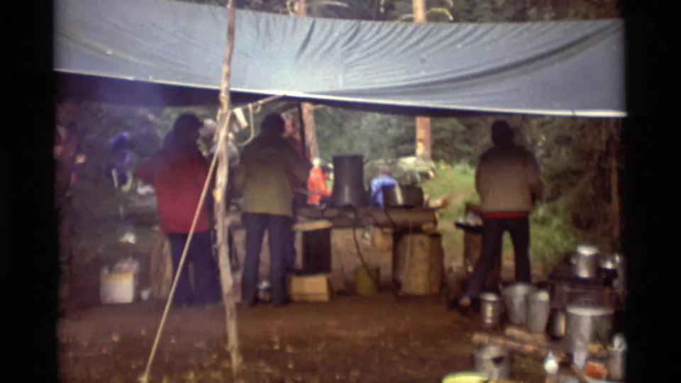 1975 Agate Springs, Montana home-movie frame showing hikers in jackets gathered under a tarp beside cookware and camp supplies.