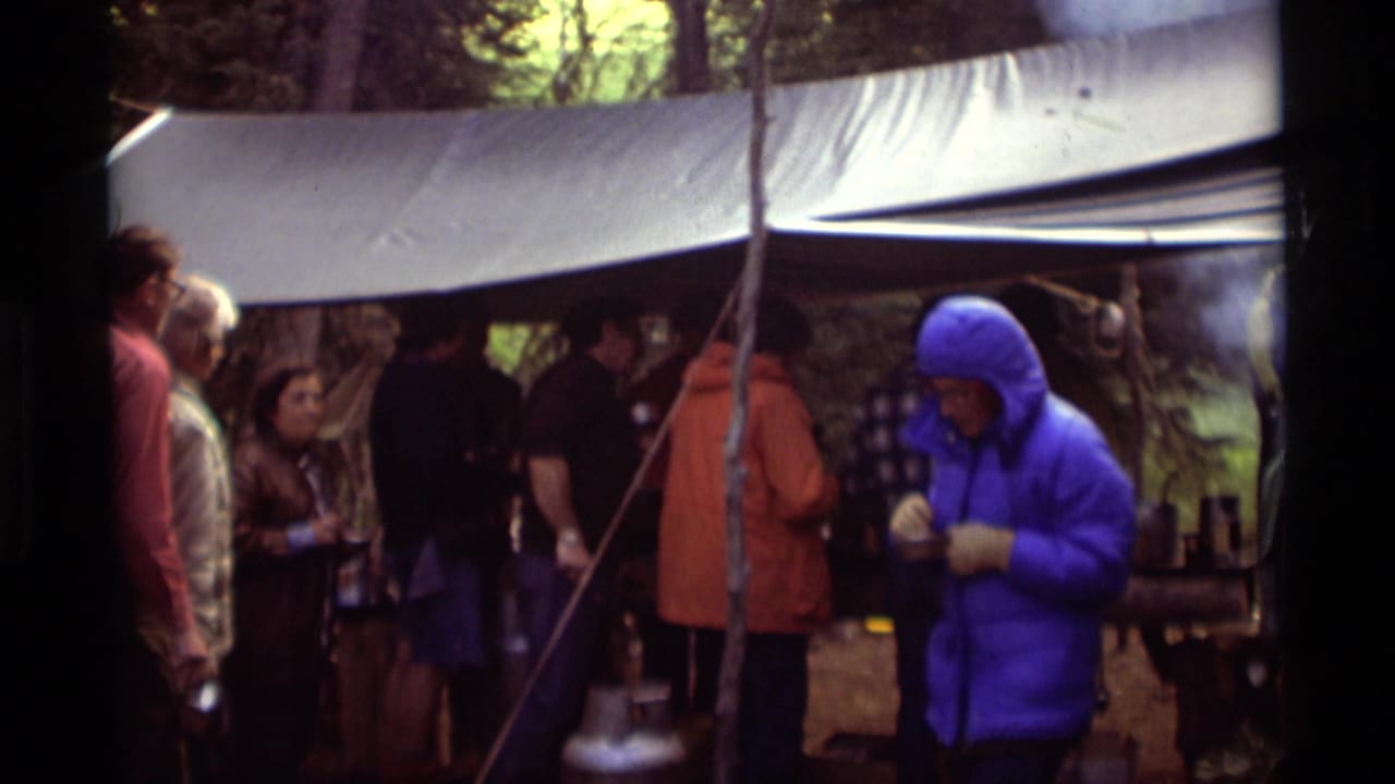 Group of adults gathers under a tarp while a person in a bright blue jacket eats nearby.