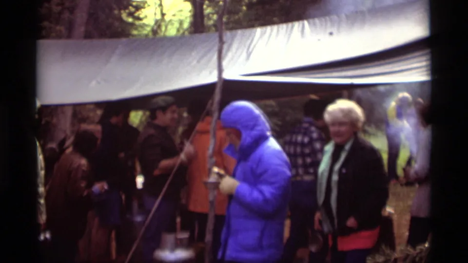1975 Montana camping frame with a bright blue jacketed person, older adult, and other hikers gathered near smoky camp cooking gear.