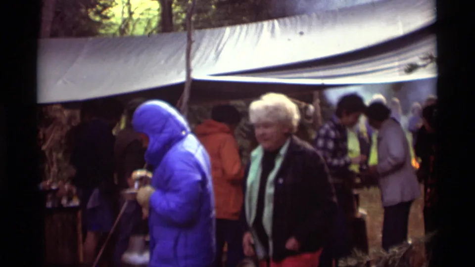 Agate Springs, Montana home-movie frame showing campers near a tarp shelter, including a blue jacketed person and an older adult in front.