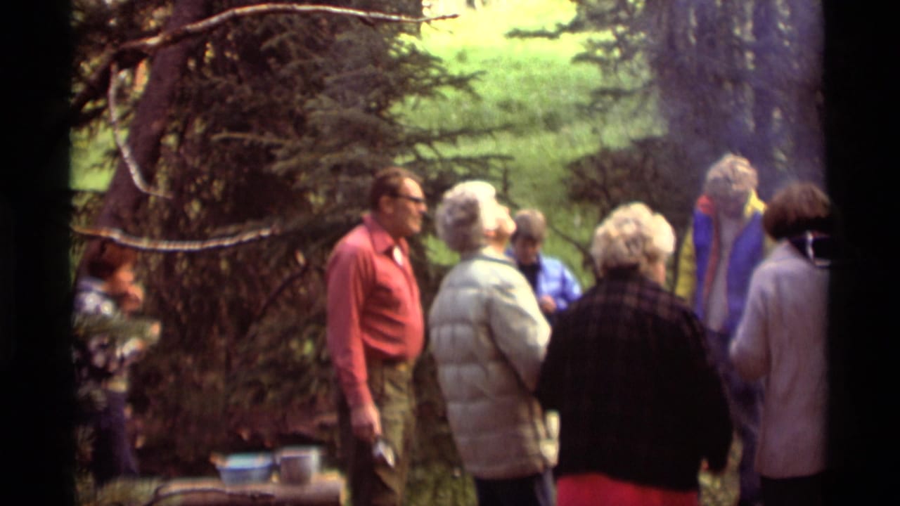 Adults in 1970s outdoor clothing converse in a wooded Agate Springs setting, with smoke drifting near cooking pots.