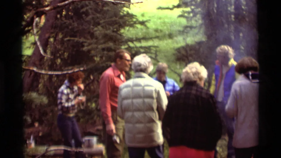 1975 Montana home-movie frame showing adults gathered in the woods near a smoky campfire and cooking area.