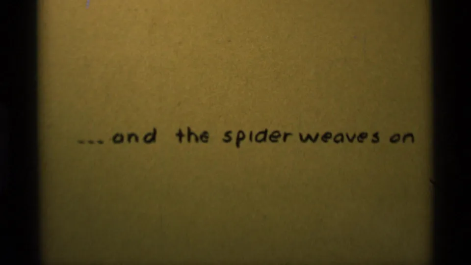 1975 8mm frame showing a handwritten phrase on aged paper: '...and the spider weaves on'.