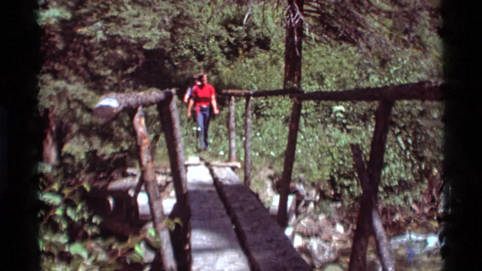 1975 Agate Springs, Montana 8mm frame showing a person crossing a rustic wooden footbridge amid forest vegetation.