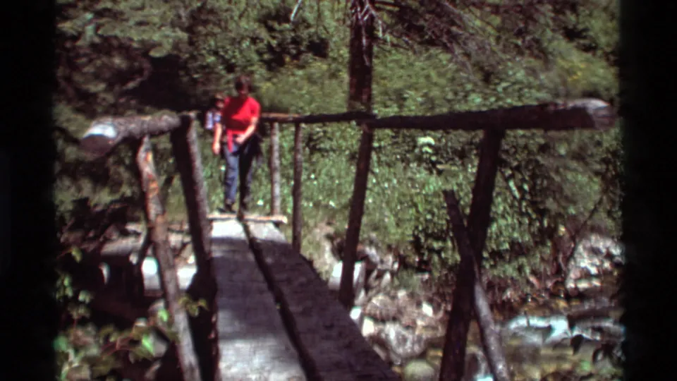 A woman in a red shirt walks onto a rustic bridge while another person stands behind her in 1975 Agate Springs, Montana footage.