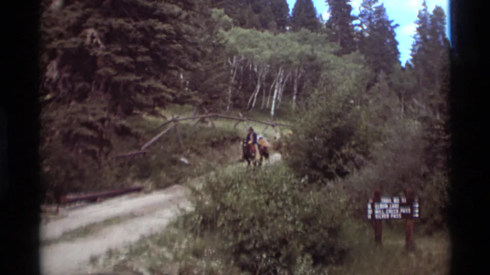 Horseback riders in the Agate Springs, Montana forest pass behind brush near a wooden trail sign in this 1975 8mm frame.