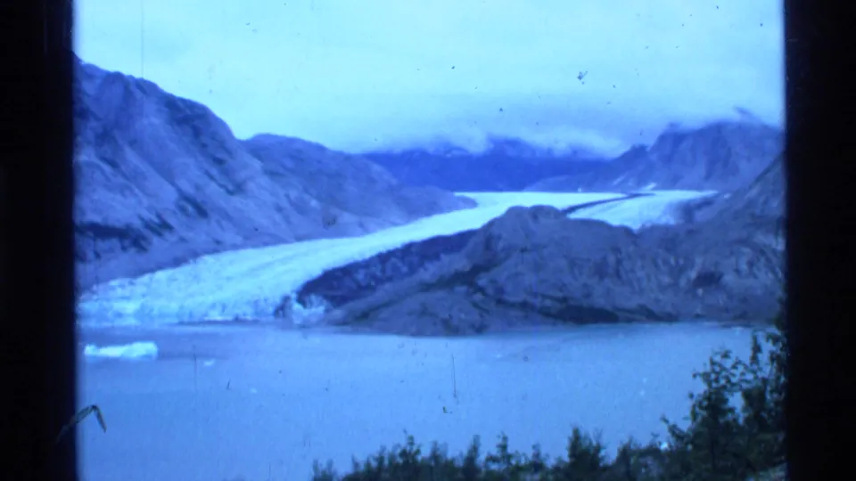 1977 Alaska 8mm view of a broad glacier reaching a calm body of water, with mountains and low clouds beyond.